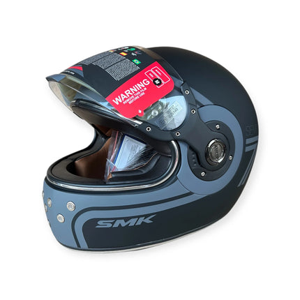 Matte black SMK full-face motorcycle helmet with clear visor and warning label